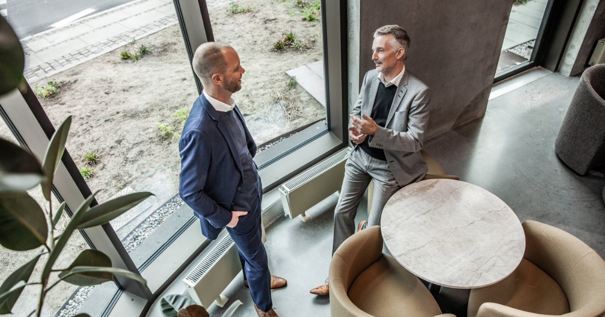 Two men are standing and talking by a window.